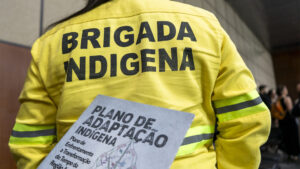 Esta imagem é um registro que capta, em primeiro plano, uma pessoa de costas vestindo um casaco de segurança amarelo-vivo com as palavras "BRIGADA INDÍGENA" estampadas em letras grandes e pretas na parte superior das costas. A pessoa está segurando um livreto ou documento com a capa voltada para cima, onde se pode ler em destaque o título: "PLANO DE ADAPTAÇÃO INDÍGENA". O subtítulo, menos visível, parece ser: "Plano de Enfrentamento e Transformação do Tempo da Região Amazônia".