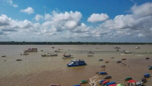 A imagem, capturada de cima (drone slide), mostra uma barqueata no que parece ser um rio largo e barrento típico da Amazônia, sob um céu parcialmente nublado, com grandes nuvens brancas em contraste com o azul. Uma grande concentração de embarcações de diversos tamanhos e cores está reunida na água, indicando um evento ou manifestação. No centro, destaca-se um barco maior, de cor azul com parte superior branca, que parece transportar um número significativo de pessoas, provavelmente os participantes do evento. Ao redor, há dezenas de barcos menores, muitos deles com coberturas coloridas (vermelhas, azuis, verdes e amarelas), que se aglomeram, principalmente na parte inferior direita da foto. Essas embarcações menores são características do transporte fluvial amazônico. Alguns barcos de lazer ou de porte médio (como iates e lanchas) também estão visíveis. A margem oposta do rio, na linha do horizonte, é coberta por uma densa vegetação florestal, reforçando o cenário amazônico. A foto transmite a sensação de um grande movimento popular e de uma celebração/protesto em massa na água, como parte das atividades da Cúpula dos Povos, destacando a importância dos rios como vias de comunicação e espaço de manifestação na região.