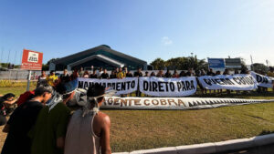 A foto mostra um grupo de pessoas reunidas em frente ao edifício de arquitetura moderna Hangar Centro de Convenções, em Belém, no Pará. No primeiro plano, à esquerda, vários indivíduos — alguns vestindo trajes tradicionais indígenas com cocares e adereços — estão agachados ou em pé, voltados para a cena central, em gestos que sugerem preparação ou reflexão antes da manifestação. O centro da foto é dominado por uma grande faixa horizontal, sustentada por um grupo diversificado de pessoas que se estende ao longo da frente do gramado. Os dizeres na faixa, em letras grandes e pretas, são claramente de protesto. A leitura parcial visível da faixa principal é “A gente cobra”.