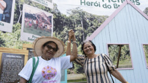 Duas mulheres extrativistas aparecem lado a lado, sorrindo e de mãos dadas, com os braços erguidos em sinal de celebração ou união. A mulher à esquerda usa um chapéu de palha, óculos e uma camiseta branca com uma estampa colorida que diz: "Somos Mulheres". A mulher à direita veste uma blusa listrada em tons de bege e preto.