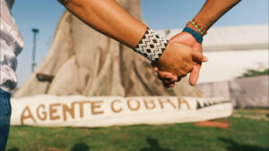 A imagem em plano médio exibe, em primeiro plano, duas mãos entrelaçadas em um gesto de união. As pessoas usam pulseiras largas (braceletes) de miçangas que remetem ao artesanato tradicional da região: uma pulseira com padrão geométrico em preto e branco, e outra mais fina com cores azul, vermelho e verde. Ao fundo, destaca-se o tronco maciço de uma grande árvore, possivelmente uma samaumeira, em um dia de sol forte. No chão, no plano inferior, uma estrutura alongada exibe a pintura: "AGENTE COBRA". A imagem articula temas de união, identidade cultural e denúncia/cobrança social, sendo um registro fotográfico de um contexto de ativismo da juventude amazônida.