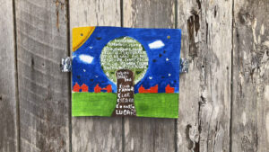 Uma pequena tela de pintura artesanal, fixada em uma parede de madeira rústica, exibe um desenho vibrante da Vila da Barca. O céu é azul escuro, com nuvens brancas, e um sol amarelo no canto superior esquerdo. No centro, uma grande árvore verde está rodeada por casas vermelhas típicas da comunidade. A tela contém mensagens de resistência e senso de pertencimento, como "VILA DA BARCA BOA" e "E NOSSO LUGAR!". No tronco da árvore e no centro, há palavras pintadas que refletem a consciência ambiental e social da Vila da Barca: "SUSTENTABILIDADE", "PLANTAR, AMAR, REDUZIR, CULTURA, RECICLAR, MAR, RIO, RESPEITAR, CONSCIÊNCIA, ATITUDE, MORAR, COMUNICAR, LUGAR". A obra simboliza a luta e o orgulho dos moradores ribeirinhos de Belém.