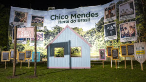 A fotografia em plano noturno, com iluminação focal, exibe uma instalação artística e memorial dedicada a Chico Mendes, ambientada em um espaço aberto, conforme sugere o nome do arquivo. No centro da imagem, destacando-se sobre um fundo de vegetação nativa da Amazônia (uma floresta), há um grande banner horizontal que proclama a figura do seringueiro e ativista. O título central, em letras grandes, é "Chico Mendes Herói do Brasil".