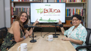 A fotografia mostra um ambiente interno, que parece ser um estúdio de gravação ou mesa de podcast/entrevista. Duas mulheres estão sentadas em frente a uma mesa de madeira clara, ambas sorrindo e apontando para um monitor de TV centralizado logo atrás delas. A mulher à esquerda tem cabelos longos e castanhos, veste uma blusa estampada escura e olha diretamente para a câmera. A mulher à direita tem cabelos curtos e cacheados, usa óculos de aros claros e uma camisa estampada em tons de verde e branco, e também olha para a câmera. Entre elas, sobre a mesa, há dois microfones de estúdio e duas canecas brancas com a logomarca do programa, além de um celular entre os microfones. O foco principal é o monitor ao fundo, que exibe a logomarca do programa: "TÔ DE BUTUCA NA COP30" em letras coloridas, com a assinatura "COM KATIA BRASIL E CRISTINA SERRA". Abaixo do título, há uma ilustração simplificada de duas pessoas em uma canoa. No topo da tela, está discretamente a logomarca da Amazônia Real.